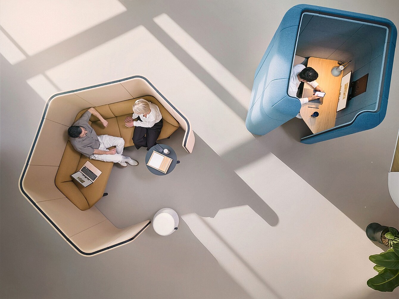 Deux personnes assises dans une cabine de bureau insonorisée beige avec une petite table et un ordinateur portable, face à une personne travaillant seule dans une cabine de bureau insonorisée bleue avec un bureau et un ordinateur portable