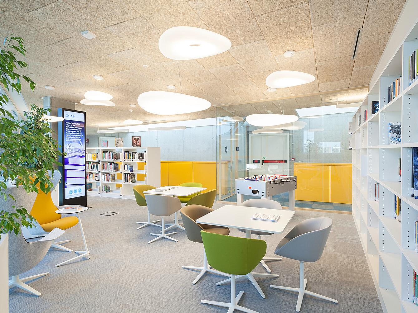 Area ufficio open space con tavoli bianchi circondati da sedie girevoli verdi e grigie, librerie bianche, mobili contenitori gialli, un biliardino e grandi lampade a soffitto