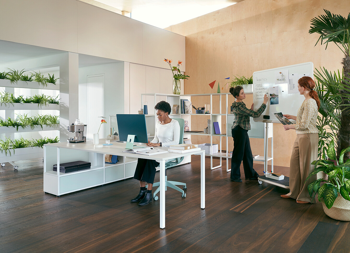 Trois femmes dans un bureau moderne agrémenté de plantes : l’une est assise à un bureau et travaille sur un ordinateur, tandis que les deux autres se tiennent debout près d’un tableau blanc et discutent de leurs notes