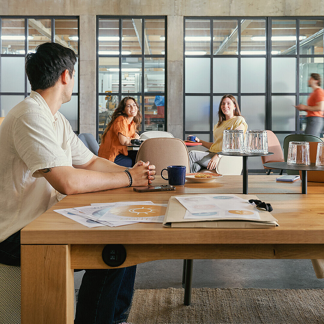 People working and talking in a modern open office with large windows and wooden tables