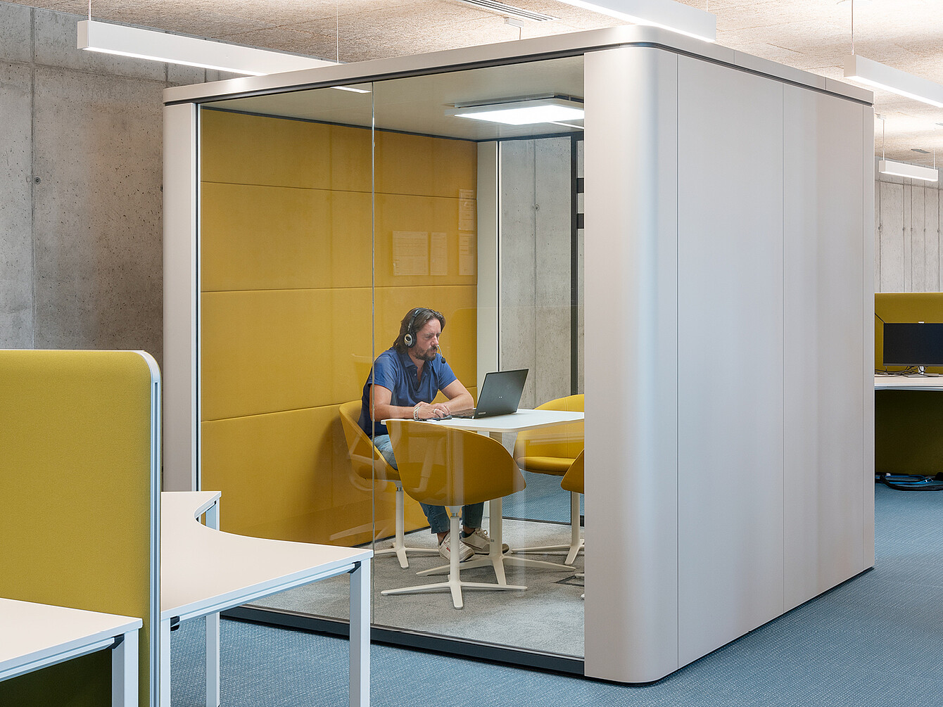 Persona con le cuffie seduta a un tavolo con un laptop all’interno di un cubicolo con pareti in vetro, pareti imbottite di giallo e sedie