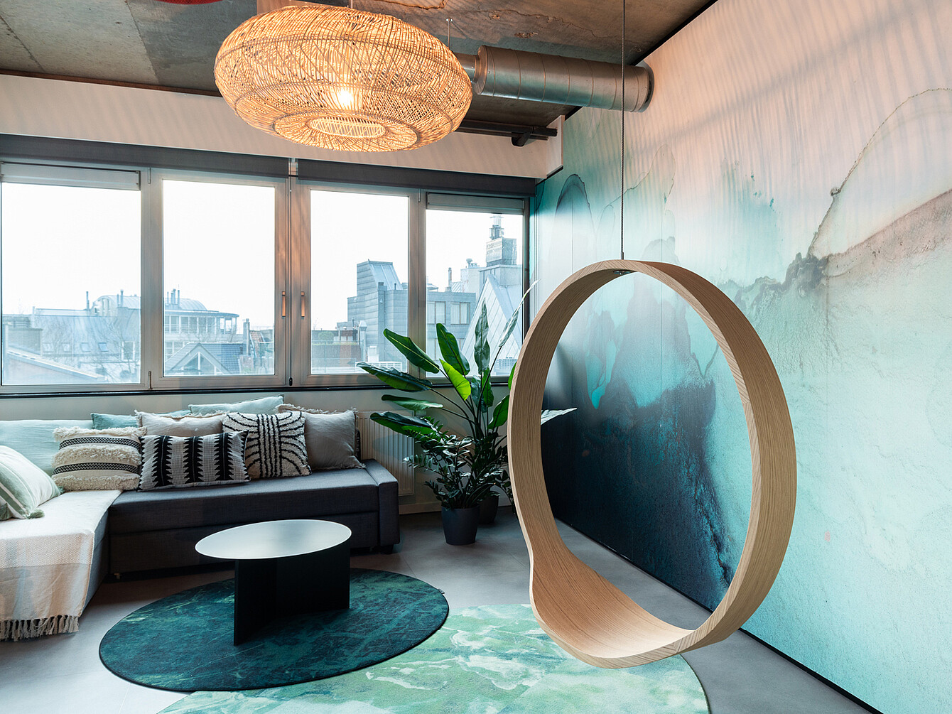 Moderna sala de estar de oficina con un sofá modular gris, cojines estampados, una mesa de centro redonda negra sobre una alfombra verde, una planta grande, una silla colgante circular de madera y un gran ventanal con vistas a los edificios de la ciudad