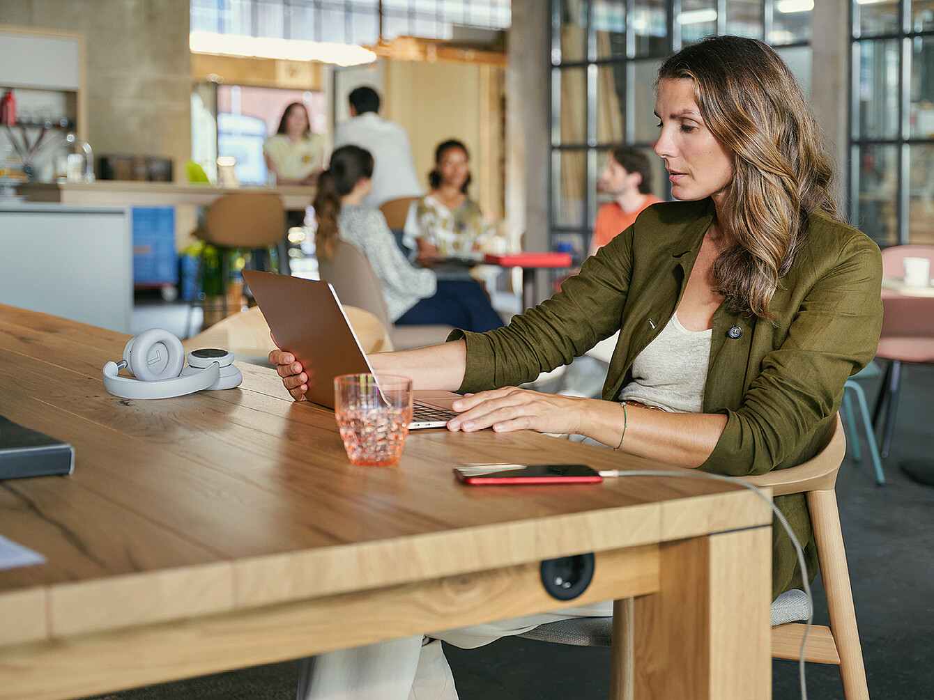 Woman sitting at wooden table in a Work Café office space, using a tablet with headphones and glass nearby, other people socializing in background