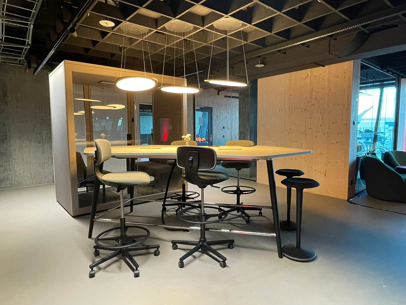 Table haute entourée de plusieurs tabourets pivotants réglables dans un espace de bureau moderne du Novu Campus, à l’aéroport de Zurich, avec des suspensions lumineuses au plafond et des murs en bois.