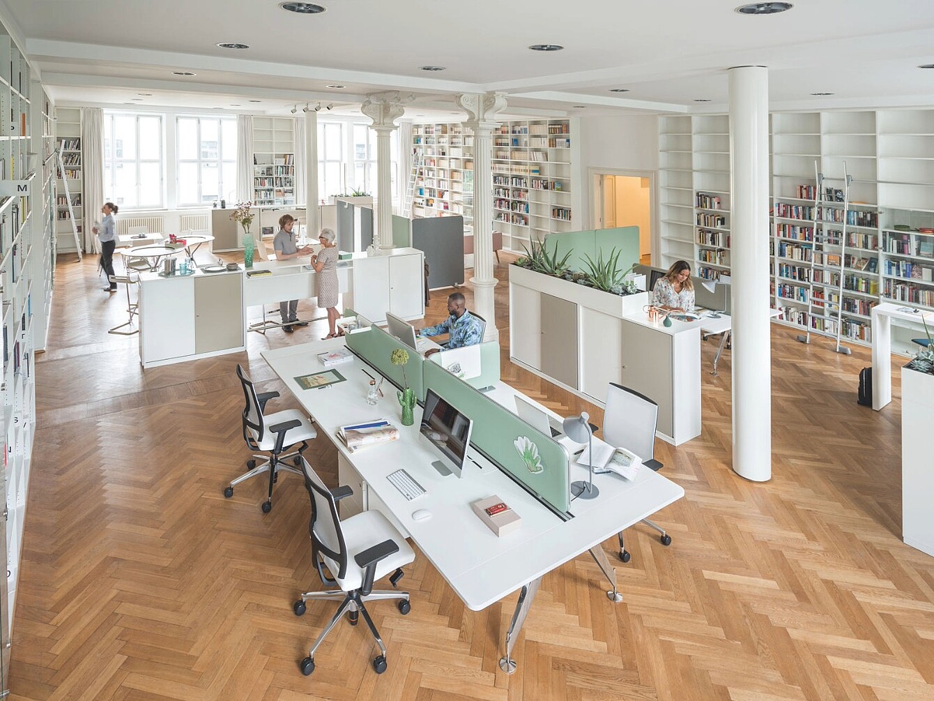 Bureau en open space avec des bureaux séparés par des panneaux en tissu vert, plusieurs personnes au travail, des étagères le long des murs et un parquet en bois