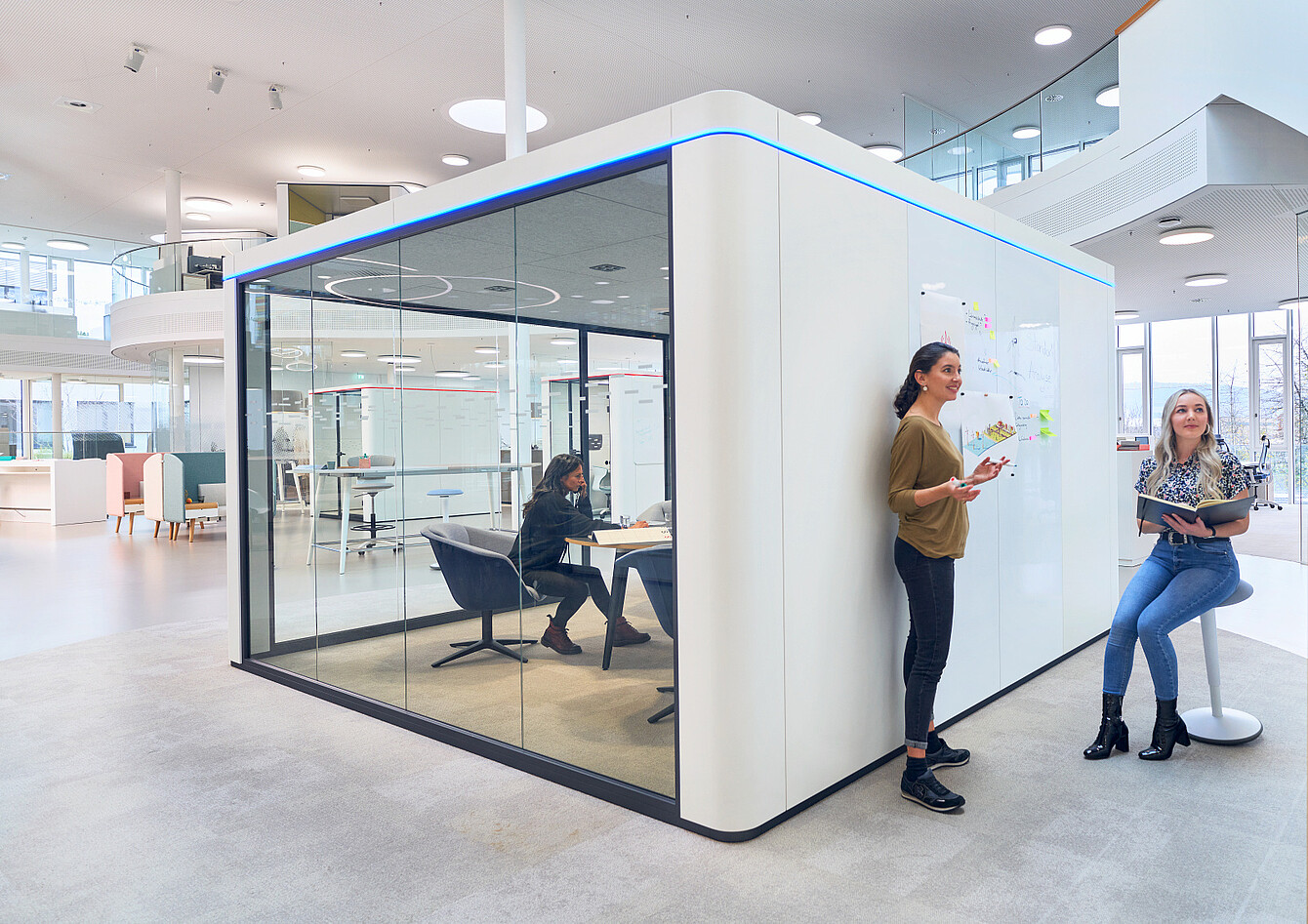 Espace de bureau moderne avec une salle de réunion aux parois vitrées et deux femmes debout et assises près d’un mur de tableau blanc couvert de notes
