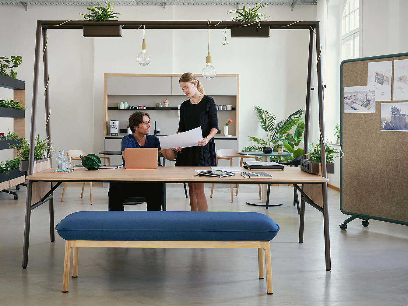 Two colleagues discussing documents in a bright Work Café office space with plants, pendant lights, and a bulletin board with architectural drawings