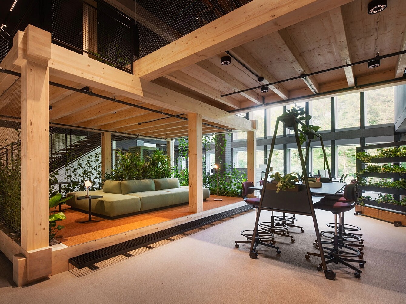 Espace de bureau ouvert avec poutres en bois, canapé vert, plusieurs chaises autour d’une table et plantes près de grandes fenêtres.