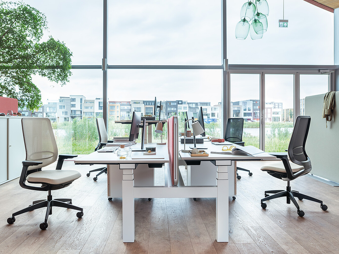 Espace de travail moderne avec quatre bureaux disposés en deux rangées face à face, des sièges ergonomiques, des écrans d’ordinateur et de grandes fenêtres offrant une vue sur la ville...