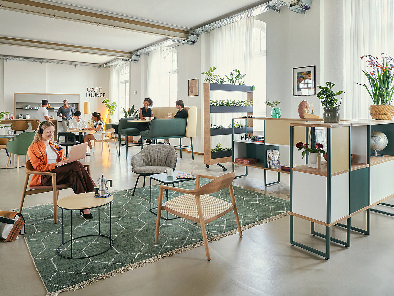 Un moderno Work Café con varias zonas de descanso decoradas con plantas. En primer plano, una mujer con una chaqueta naranja está sentada en una silla con un ordenador portátil. Al fondo, hay personas conversando y trabajando en las mesas. El espacio incluye estanterías con objetos decorativos y un cartel que dice «CAFE LOUNGE».