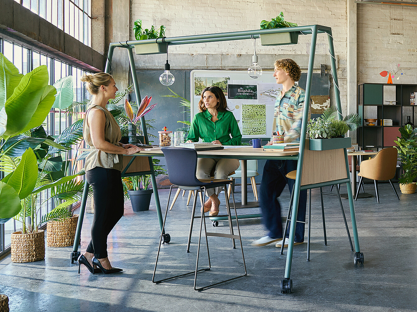 Tre colleghi collaborano attorno a un tavolo alto con piante sopra in un ufficio luminoso e pieno di verde, esemplificando il design biofilico del Garden Work Café.