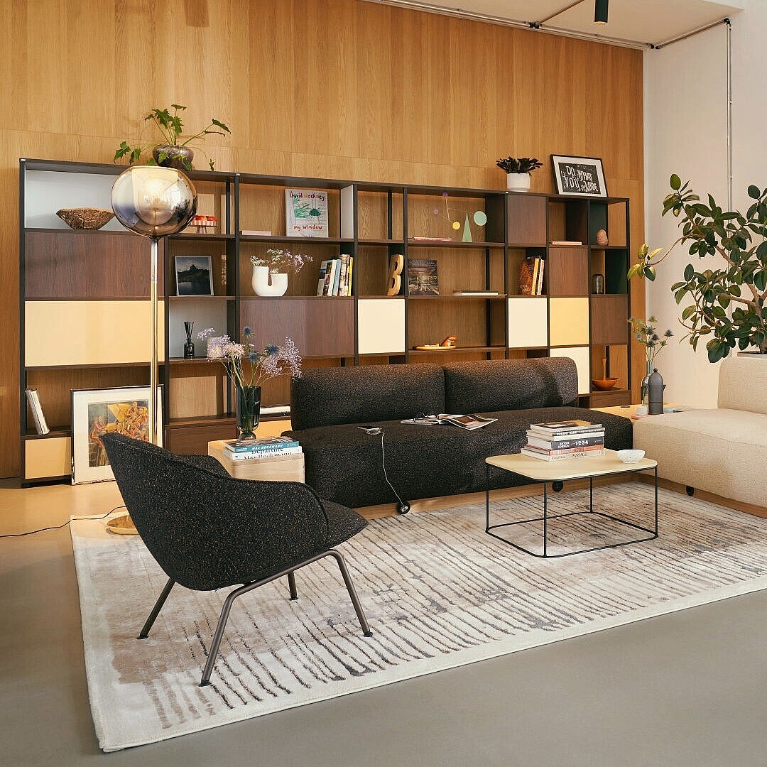 Durchdachte New Work Lounge mit zwei Sofas, Sesseln, Teppich und großem Regal vor Holzpaneelen, angrenzend an hellen Besprechungsraum mit Glaswand